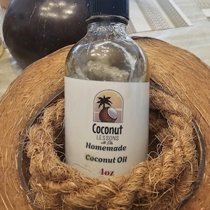 Puede incluir: Una botella de vidrio transparente con tapa negra, etiquetada como "Coconut Lessons with Ela Homemade Coconut Oil 4oz." La botella está dentro de una cáscara de coco tejida, con una cáscara de coco marrón oscuro en el fondo.