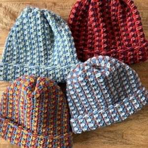 May include: Four handmade crochet beanies in various colors. The beanies are blue, red, and a mix of blue, yellow, and red. The hats have a textured, woven appearance and are displayed on a wooden surface.