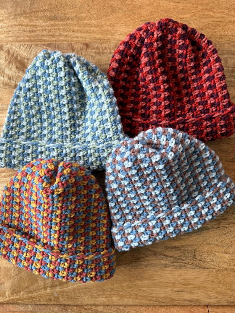 May include: Four handmade crochet beanies in various colors. The beanies are blue, red, and a mix of blue, yellow, and red. The hats have a textured, woven appearance and are displayed on a wooden surface.