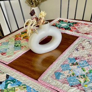 May include: A collection of colorful, quilted placemats with a patchwork design, featuring various floral and geometric patterns. Each placemat has a pink striped border. A white, donut-shaped vase holds dried flowers on the wooden table.