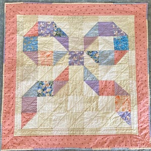 May include: A handmade quilt with a cream-colored center and a pink border. The quilt features geometric patterns in pastel colors, including blue, purple, and peach, with floral and patterned fabric accents. The quilt is approximately 50 inches square.