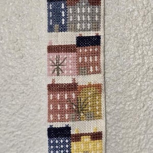 Könnte beinhalten: Ein weißer Wandbehang aus Stoff mit einem Kreuzstich-Design aus bunten Häusern und Bäumen. Der Behang hat einen Messinghaken und eine Glocke unten.