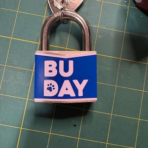 May include: A silver padlock with a blue label that reads "BU DAY" in white block letters, with a paw print replacing the "O". The padlock is attached to a set of keys. The background is a green cutting mat.