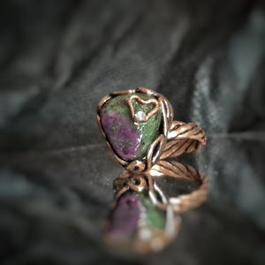 May include: A copper ring featuring a teardrop-shaped stone. The stone is a blend of green and purple, with a small diamond and a heart-shaped detail. The ring's band has a twisted rope design, with a mirrored reflection.