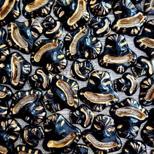 May include: A collection of small, black ceramic mask-shaped ornaments featuring gold-painted accents on the facial features and edges. Each piece has a hollowed-out mouth area and is arranged in a dense, overlapping pattern across the frame.