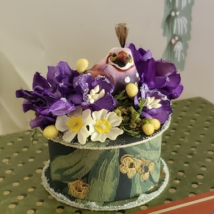 May include: A decorative round box with a bird figurine perched atop a bed of purple and white flowers. The box is dark green with a leaf pattern and gold accents. The bird has a brown and purple body.