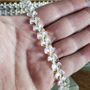 May include: White and gold floral trim. The trim features a repeating pattern of white fabric flowers with gold-colored metal accents. The trim is held in a hand, with more trim visible in the background.