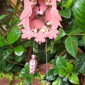 May include: A pink wooden birdhouse-shaped ornament with a clock face, birds, and floral accents. Two pink pinecone-shaped ornaments hang below on silver chains. The ornament is surrounded by green foliage.