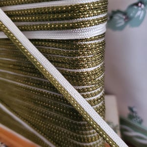 May include: Close-up of a spool of decorative trim. The trim features a combination of olive green and gold metallic braided cord, with white cord accents. The trim is likely used for sewing, crafting, or embellishing various items.