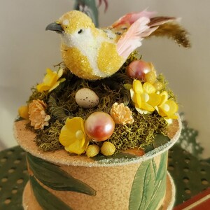 May include: A small, round, decorative box with a yellow bird perched on top. The box is covered in green leaves and has a brown base. The bird has pink wings and a yellow body with white accents. The box is decorated with yellow flowers, green moss, and small white eggs.