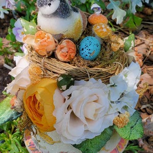 May include: A decorative bird figurine sits atop a floral arrangement. The arrangement includes a nest with speckled eggs, orange and yellow flowers, and a base with a painted design. The bird has a black and white head and a yellow and brown body.