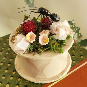 May include: A small, round, pink box with a white trim and a glittery finish. The box is decorated with a miniature ant, pink flowers, green moss, and red berries.