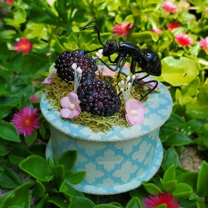 May include: A decorative box with a light blue and white pattern, topped with faux blackberries, pink flowers, and a black ant figurine. The box is surrounded by green foliage and small pink flowers.