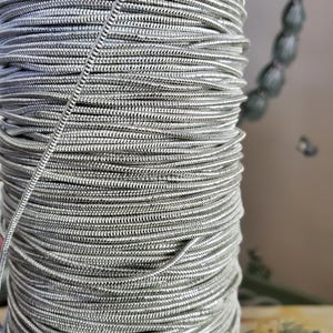 May include: Close-up of a spool of silver-colored, flexible cord. The cord is tightly wound, creating a cylindrical shape. The texture of the cord appears to be ribbed or woven, reflecting light. The cord is likely used for jewelry making or crafts.
