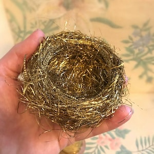May include: A gold-colored bird's nest made of shiny metallic strands. The nest is held in a person's hand.
