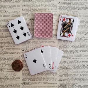 May include: A collection of playing cards including the six of spades, jack of diamonds, ace of clubs, and two of clubs, alongside a copper penny, all arranged on a page of a dictionary with visible text definitions and columns of printed words.
