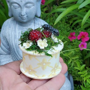 May include: A small, round, decorative box with a floral design in yellow and white. The top is adorned with red berries, green moss, white flowers, and a black insect figurine. The box is held in front of a gray Buddha statue.