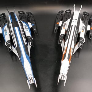 Puede incluir: Dos naves espaciales modelo, una blanca con detalles naranjas y la otra azul con detalles negros. Ambas naves tienen el texto "NORMANDY" y "SR2" impreso en ellas.