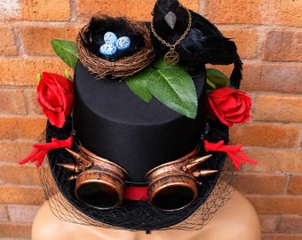 May include: A black top hat adorned with steampunk goggles, red roses, and a black crow wearing a gold pendant. A nest with blue speckled eggs sits on top. A black veil and red accents.