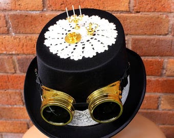May include: A black top hat with gold-rimmed goggles and a white lace doily on top. The hat is decorated with miniature gold tea party accessories. The goggles are attached with a black strap.