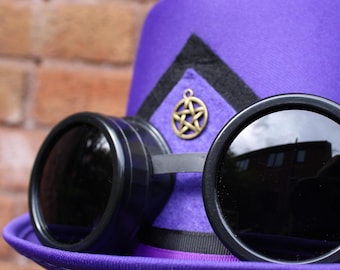May include: A vibrant purple top hat with a black trim and a gold-coloured emblem, complemented by black steampunk goggles. The hat features a broad brim and a decorative band. The goggles have circular lenses.