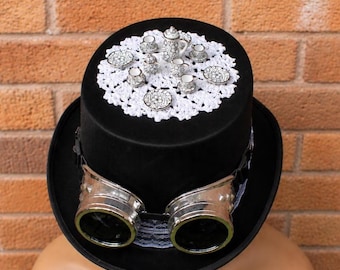 May include: A black top hat featuring silver goggles and a white lace doily. The hat is decorated with a miniature silver tea set. The background is a red brick wall.