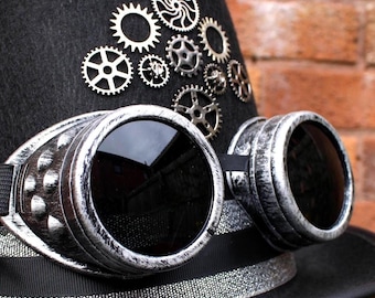 May include: A black steampunk top hat with silver-rimmed goggles and decorative gears. The hat has a black ribbon and silver trim. The goggles have dark lenses and a black strap. The gears are in various sizes and designs.