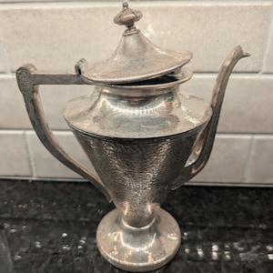 May include: A silver-toned metal teapot featuring a hammered texture on the body, a conical base, a curved handle, a long spout, and a hinged lid topped with a small finial. The surface has a metallic sheen with visible indentations.