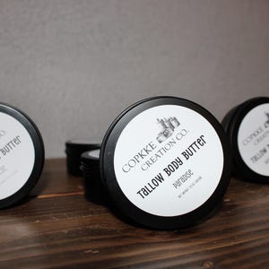 Könnte beinhalten: Mehrere schwarze Dosen Talg-Körperbutter sind auf einer Holzoberfläche angeordnet. Jede Dose hat ein weißes Etikett mit schwarzem Text, auf dem "Tallow Body Butter" und der Markenname stehen. Die Etiketten enthalten auch den Produktnamen.