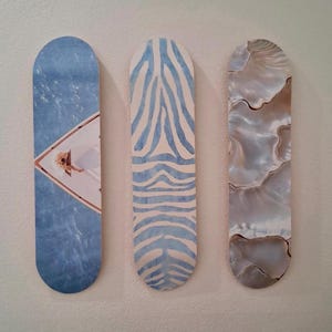 May include: Three decorative wall art pieces. The first is blue with a diamond-shaped image. The second has a blue and white zebra stripe pattern. The third has a pearlescent, marbled design in shades of gray and brown.