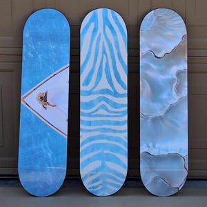 May include: Three skateboards with unique designs. The first features a blue water scene with a person on a boat. The second has a blue and white zebra stripe pattern. The third has a pearlescent, marbled effect.