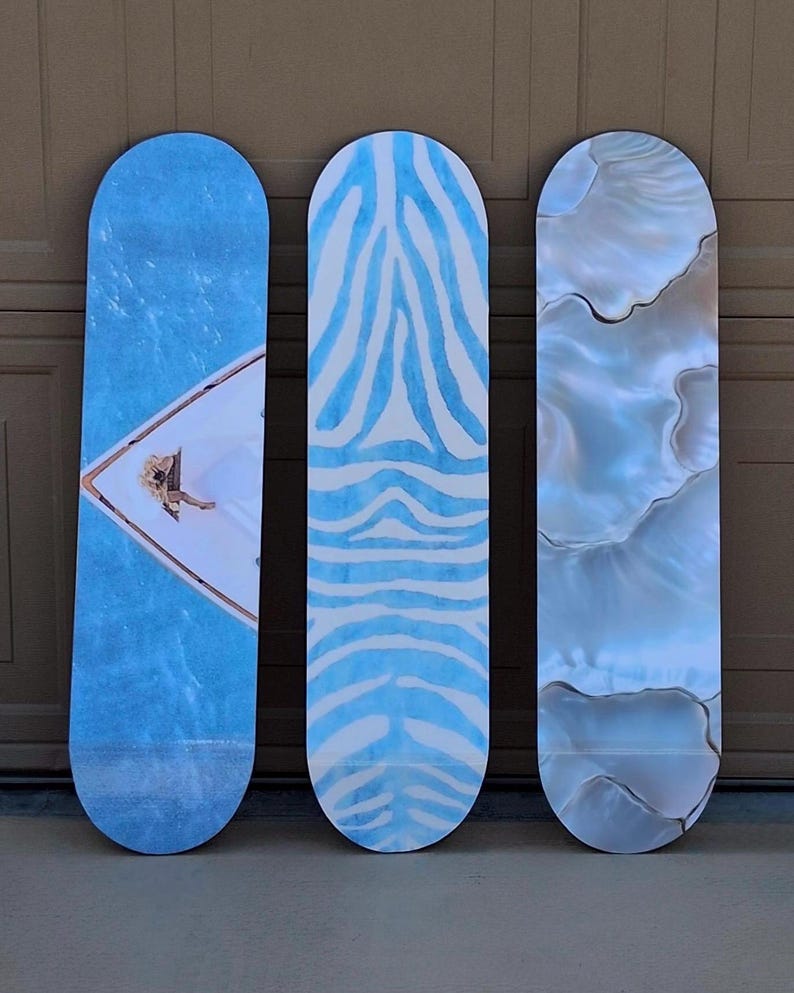 May include: Three skateboards with unique designs. The first features a blue water scene with a person on a boat. The second has a blue and white zebra stripe pattern. The third has a pearlescent, marbled effect.