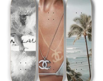 May include: Set of three skateboards. One features a black and white image of a surfer in the ocean. The second shows a person's chest with a silver necklace and a logo. The third displays a beach scene with palm trees and ocean.
