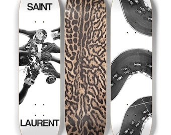 May include: Three skateboards with unique designs. One features a black and white image of hands holding glasses, with "SAINT LAURENT" text. Another has a leopard print pattern. The third displays a race track with cars.