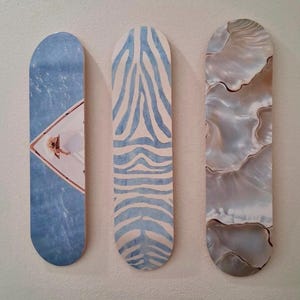 May include: Three decorative skateboard-shaped wall art pieces. The first features a blue water scene with a person, the second has a blue zebra stripe pattern, and the third has a pearlescent, shell-like design.