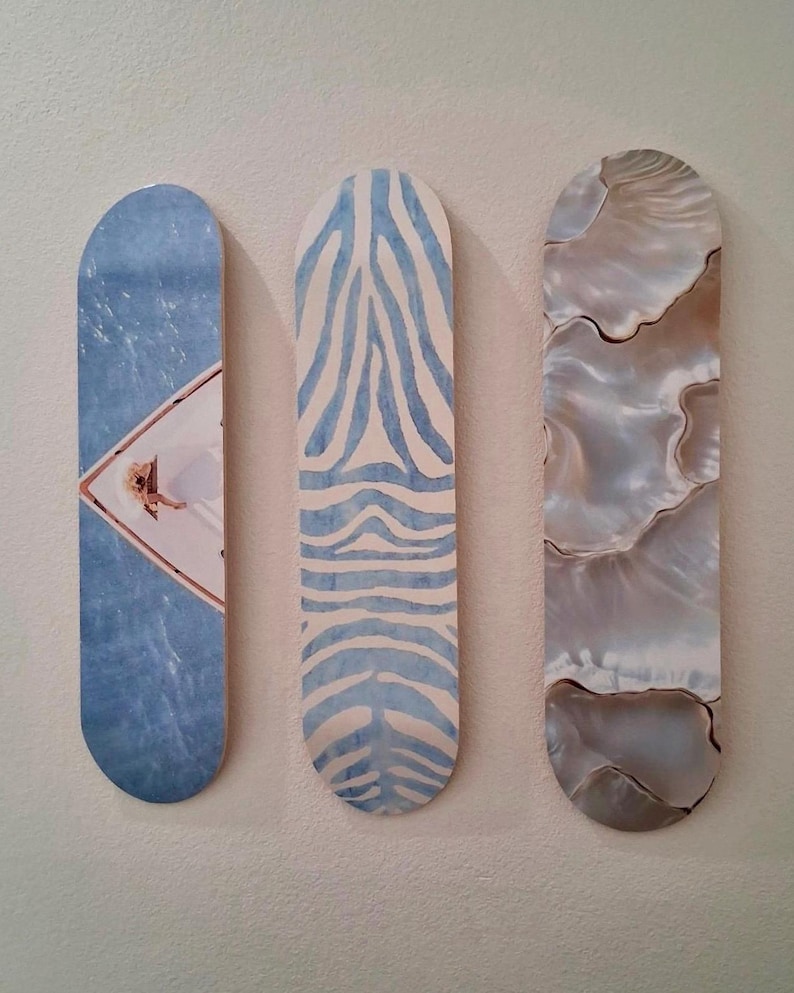 May include: Three decorative skateboard-shaped wall art pieces. The first features a blue water scene with a person, the second has a blue zebra stripe pattern, and the third has a pearlescent, shell-like design.