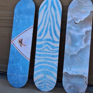 May include: Three skateboards leaning against a brown surface. The boards feature unique designs: one with a blue water-like pattern and a figure, another with blue zebra stripes on white, and the last with a pearlescent, marbled effect.