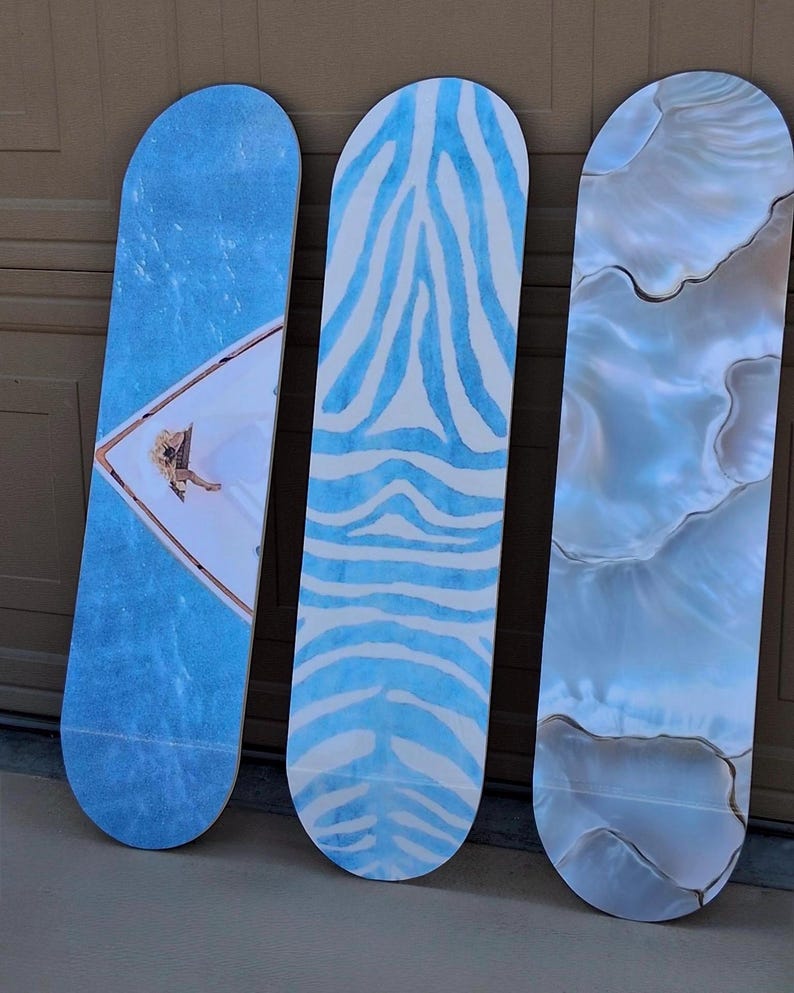 May include: Three skateboards leaning against a brown surface. The boards feature unique designs: one with a blue water-like pattern and a figure, another with blue zebra stripes on white, and the last with a pearlescent, marbled effect.