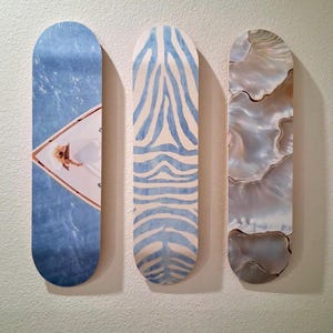 May include: Three skateboard decks with unique designs. One features a blue water scene with a person on a boat, another has a blue zebra stripe pattern, and the last has a pearlescent, shell-like design.