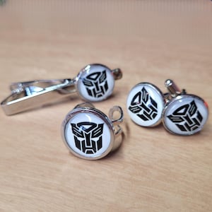 Könnte beinhalten: Eine silberfarbene Krawattenklammer und ein Paar passende Manschettenknöpfe, jeweils mit einer runden weißen Einlage und einem schwarzen geometrischen Autobot-Logo. Das Set besteht aus einer Krawattenklammer mit Federclip und zwei runden Manschettenknöpfen.