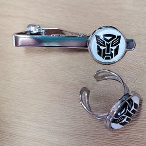 Könnte beinhalten: Eine silberfarbene Metall-Krawattenklammer und ein passender verstellbarer Ring, beide mit einer runden weißen Einlage und einem schwarzen Autobot-Symbol aus der Transformers-Reihe. Die Krawattenklammer hat einen Klappverschluss und der Ring ist offen.