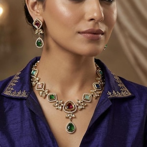 May include: A gold necklace and earring set with colourful gemstones. The necklace features a central pendant with a large green stone, and the earrings have a similar design with red and green stones.