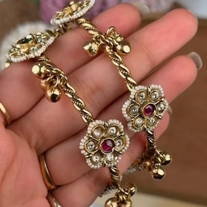 May include: Two gold-toned metal bangles featuring a twisted band design. Each bangle is adorned with floral-shaped accents containing a central red stone surrounded by small white beads. Small gold-toned spherical charms hang from the twisted bands.