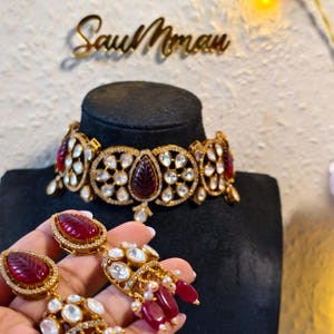 May include: A gold-coloured jewellery set comprising a choker necklace and matching earrings. The necklace and earrings feature red and clear gemstone embellishments. The necklace is displayed on a black mannequin head. The earrings are held in a person's hand. The text "Sau Mman" is visible.