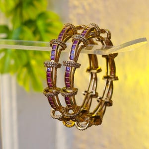 Könnte beinhalten: Goldfarbene Armreifen mit lila rechteckigen Edelsteinen und kleinen klaren Steinen. Die Armreifen werden auf einem transparenten Regal präsentiert, mit einer verschwommenen grünen Pflanze im Hintergrund. Der Schmuck hat ein verziertes Design.