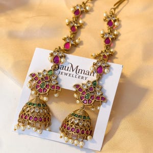 May include: A pair of gold-toned jhumka earrings featuring pink and green stones, clear crystals, and small white pearl accents. The earrings are attached to a white display card with the text "bauMman JEWELLERY" printed in black ink.