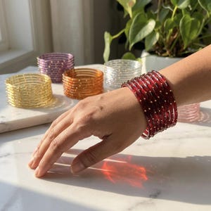 May include: A collection of thin, circular bangles in various translucent colours including red, yellow, purple, orange, and clear, each featuring small, evenly spaced metallic gold-toned studs embedded along the outer surface of the bands.