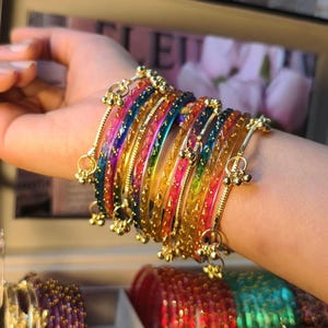 Könnte beinhalten: Eine Sammlung farbenfroher Armreifen in Blau, Grün, Gelb, Orange und Rosa. Die Armreifen sind mit goldfarbenen Akzenten und kleinen, hängenden Glöckchen verziert. Das Bild zeigt eine Vielzahl lebendiger, dekorativer Armbänder.