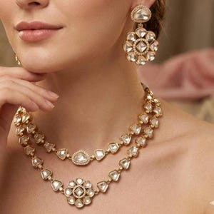 Peut inclure: Gros plan sur un ensemble de bijoux dorés comprenant un collier et des boucles d'oreilles. Le collier comporte deux rangs avec des pierres claires de forme géométrique et un pendentif floral. Les boucles d'oreilles ont un design floral similaire.