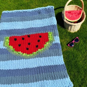 May include: A blue and gray striped blanket with a large watermelon design. A woven basket with a slice of watermelon and pink sunglasses are on the green grass.